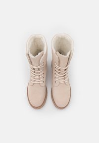 Botas de tobillo de ante beige con forro de piel sintética, suela plana marrón, y ojetes metálicos, con cordones blancos y detalles de costura en contraste.
