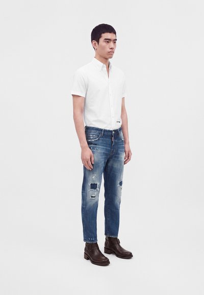 Homme portant une chemise blanche à manches courtes boutonnée, un jean bleu délavé et des bottines marron foncé, debout devant un fond uni.