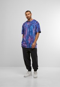 Mand står iført en stor oversize T-shirt med lilla og blå mønster, sorte joggers og hvide sneakers foran en ensfarvet grå baggrund.