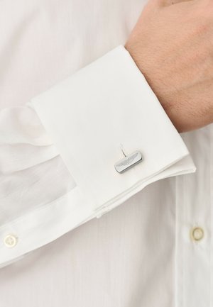 Valge triiksärgi mansett, mis on kinnitatud hõbedase ristkülikukujulise mansetinõelaga, millele on graveeritud tiivuline embleem.