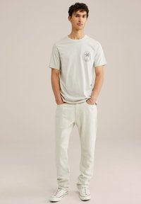 Lys grå T-shirt med rund halsudskæring og grafisk tryk af en palme på forsiden; parret med lyse beige bukser og hvide sneakers. Glat stofstruktur.
