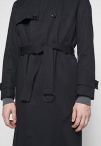 Trench-coat noir à ceinture avec boutons croisés, détail à anneau D, poches à rabat et poignets de manches gris visibles aux poignets.