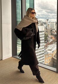 Długi brązowy płaszcz z beżowym szalem narzuconym na ramiona. Ciemna torebka i rękawiczki. Tło przedstawia miejską panoramę z szklanymi oknami.