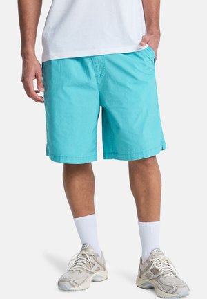 Mann trägt türkisfarbene Shorts, weiße Sportsocken, beige Reebok-Sneaker und ein weißes T-Shirt, mit einer Hand in der Tasche.