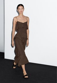 Hnědé šaty vrstvené s tenkými ramínky, s kaskádovými volány a splývavým střihem; kombinované s černými plochými sandály. Minimalistické pozadí.