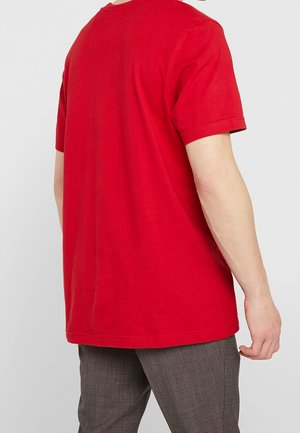 Eine Person steht mit dem Rücken zur Kamera, trägt ein leuchtend rotes T-Shirt mit kurzen Ärmeln und graue karierte Hosen vor einem schlichten, hellen Hintergrund.