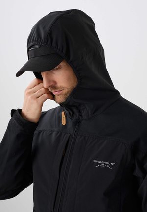 Hombre con una chaqueta negra con capucha y una gorra negra, ajustándose la capucha con la mano, con una ligera barba visible, el logo en el pecho de la chaqueta dice Swedemount.