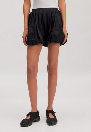 Persoon met een wit shirtje, zwarte ruime shorts met elastische taille en zoom, en zwarte schoenen met gespleten teen en riempjes.