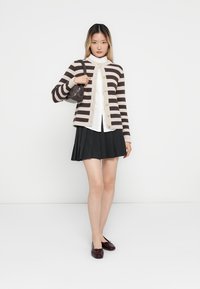 Stripet cardigan i beige og brun, hvit skjorte med krage, plissert svart skjørt, og burgunder sko med en liten brun håndveske.
