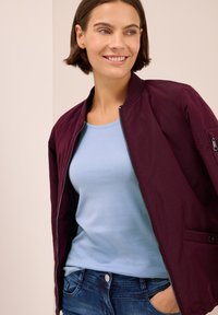Femme aux cheveux bruns courts sourit, vêtue d'une chemise bleu clair, d'un bomber violet foncé ouvert et d'un jean bleu avec une main dans la poche.
