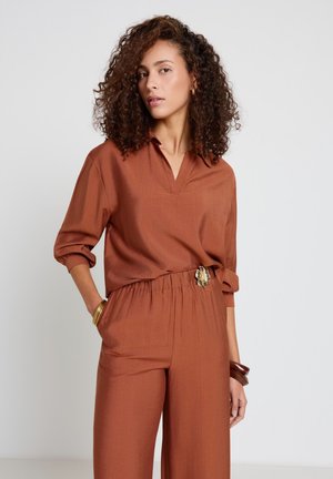 Frau mit lockigem Haar, die ein rostfarbenes Langarmoberteil und passende Hosen mit floralem Gürtelmotiv trägt, posiert mit einer Hand in der Tasche.