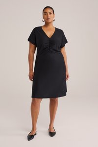 Zwarte jurk met korte mouwen en V-hals, voorzien van gatherdetails bij de buste en een losse fit, gemaakt van gladde stof. Draagt met zwarte schoenen.