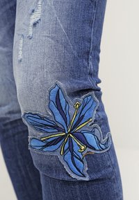 Denim jeans with a distressed blue finish, featuring a large blue floral embroidery on the left knee, accentuated by yellow details.