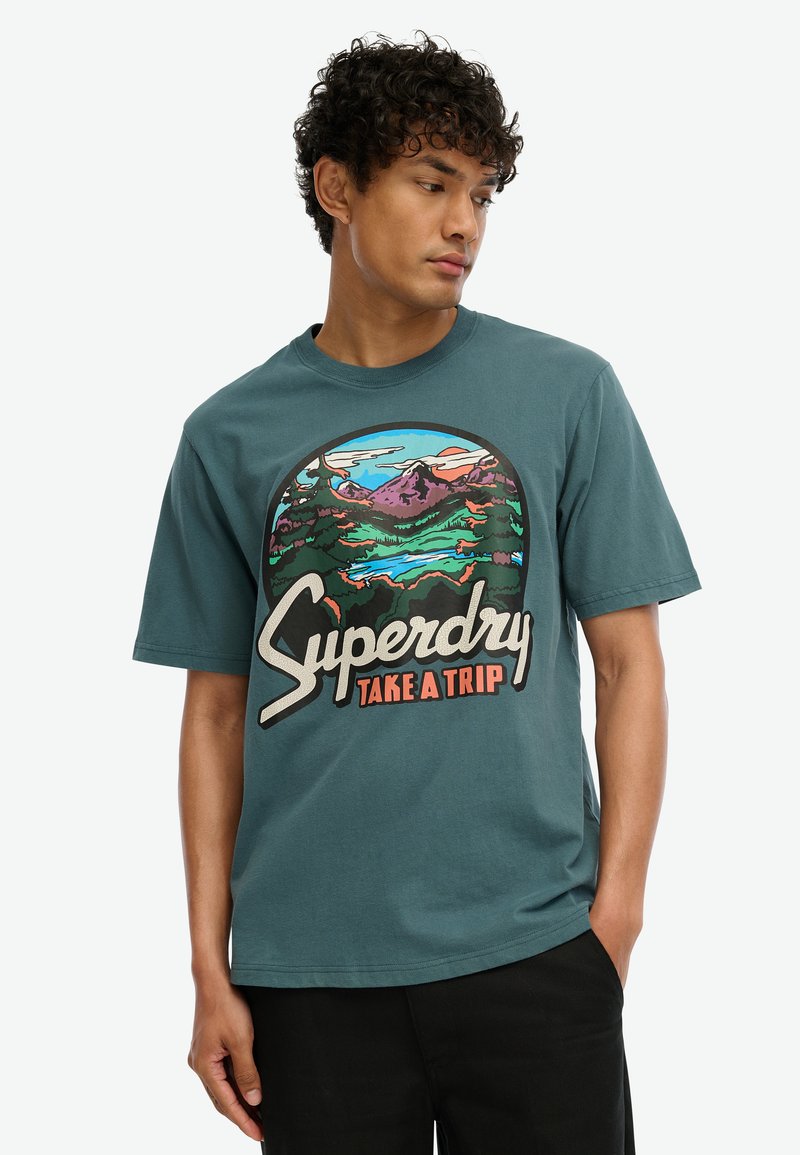 Camiseta de algodón color teal con un estampado gráfico de montañas y árboles, y el texto "Superdry Take a Trip" en colores contrastantes y en negrita.