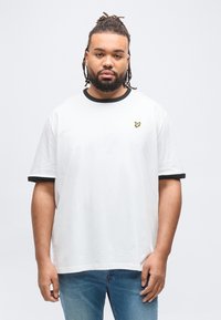 Άνδρας με γενειάδα και κοτσιδάκια, φορόντας λευκό μπλουζάκι με μαύρο περίγραμμα και μικρό κίτρινο λογότυπο πουλιού, στέκεται μπροστά σε απλό ανοιχτόχρωμο φόντο.