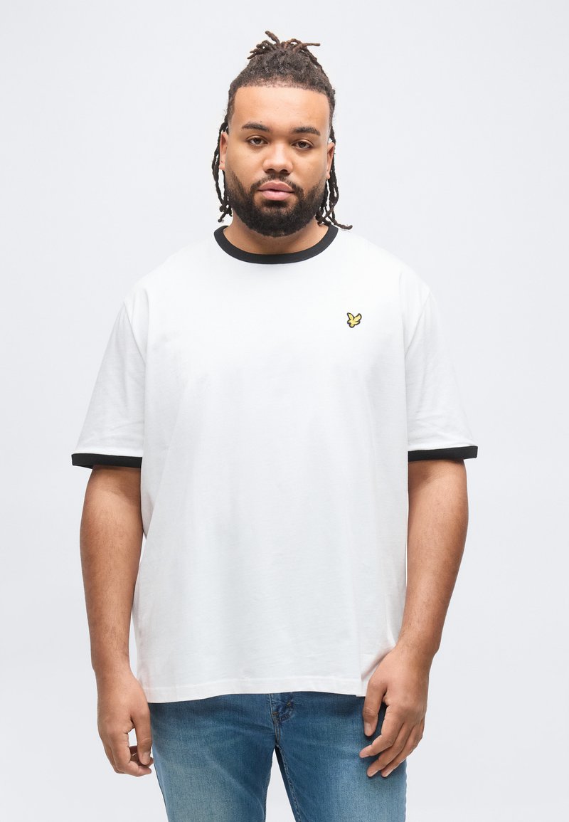 Άνδρας με γενειάδα και κοτσιδάκια, φορόντας λευκό μπλουζάκι με μαύρο περίγραμμα και μικρό κίτρινο λογότυπο πουλιού, στέκεται μπροστά σε απλό ανοιχτόχρωμο φόντο.