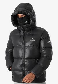 Homme portant une doudoune noire avec capuche et masque facial, affichant un logo de montagne en Helvetica sur la poitrine et la manche, les mains partiellement dans les poches.