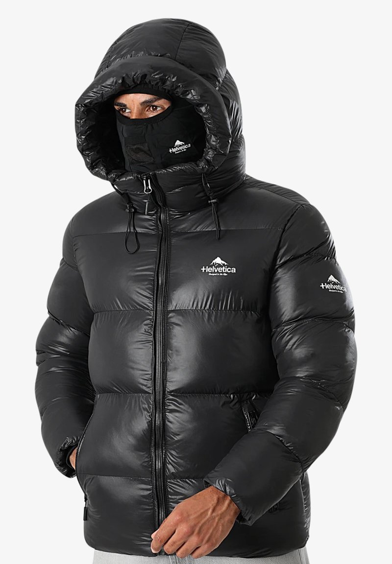 Homme portant une doudoune noire avec capuche et masque facial, affichant un logo de montagne en Helvetica sur la poitrine et la manche, les mains partiellement dans les poches.