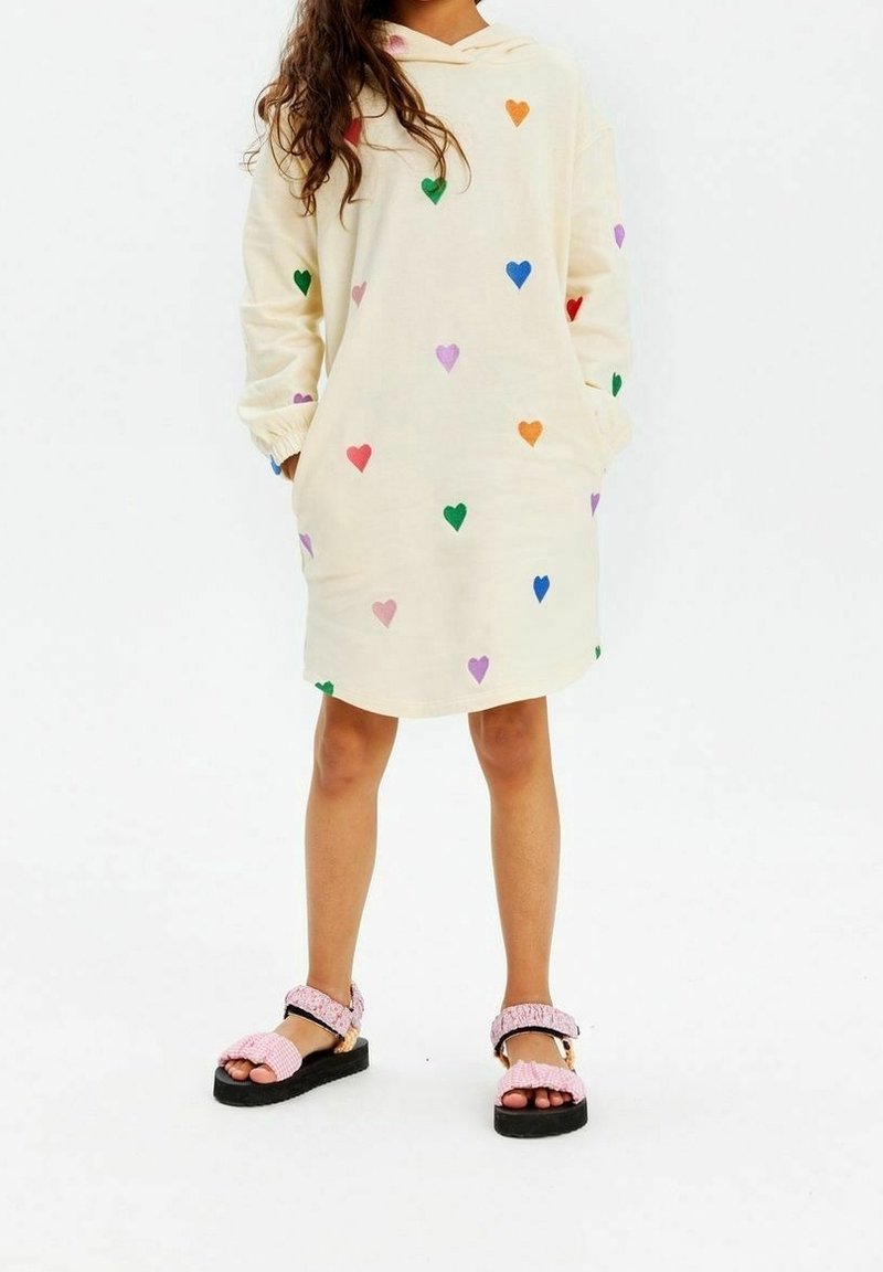 Abito con cappuccio di colore crema con motivi di cuori multicolori; abbinato a sandali rosa testurizzati con cinturini regolabili e suole nere.