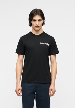 Giovane uomo con capelli scuri di media lunghezza che indossa una maglietta nera con il logo "Replay" sul petto e pantaloni blu scuro, in piedi contro uno sfondo bianco.