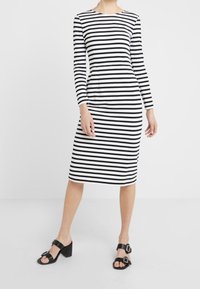 Kobieta w czarno-białej poziomo paskowanej sukience midi z długim rękawem, nosząca czarne sandały na obcasie z klamrami.