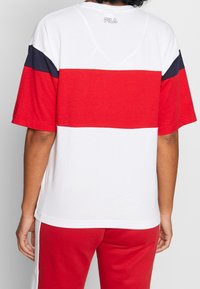 Camiseta de color bloqueado en rojo y blanco con mangas cortas, escote redondo y acentos en azul marino. Presenta un logo en la parte superior de la espalda. Tejido suave.