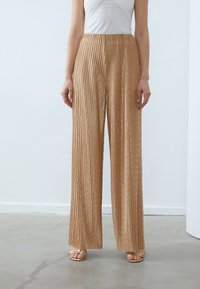 Pantalons plissés à jambes larges en or métallique avec un motif texturé, dotés d'une taille haute et d'une finition lisse. Portés avec des sandales à brides.