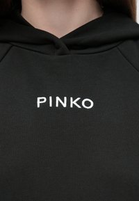 Camisola preta com capuz, apresenta um bolso canguru à frente, capuz com cordão ajustável e a marca "PINKO" bordada a branco no peito. Textura suave.