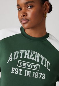 Zelená mikina s bielymi raglánovými rukávmi; na prednej strane obsahuje opotrebovaný biely text "AUTHENTIC LEVI’S EST. IN 1873", vyrobená z mäkkého bavlneného materiálu.