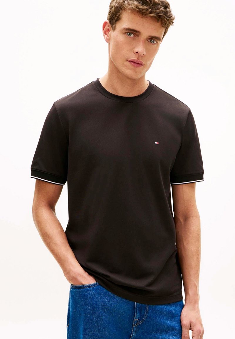 Jeune homme portant un t-shirt noir uni avec un petit logo sur la poitrine et un jean bleu, debout avec une main dans la poche devant un fond clair.