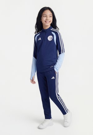 Osmijehnuta djevojka s dugom tamnom kosom koja nosi Adidas trenirku tamnoplave i svijetloplave boje te bijele tenisice, stoji s jednom rukom u džepu.