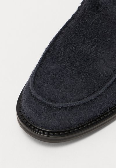 Sötétkék filcből készült, slip-on stílusú cipő, lekerekített formával, struktúrált felsőrésszel és keskeny fekete talppal, amelyen varrási részletek találhatók.