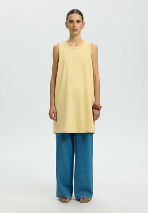 Kvinde med håret bundet tilbage bærer gul ærmeløs tunika, brede blå denimbukser, sandaler og brune armbånd, stående mod ensfarvet baggrund.