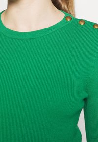 Haut en tricot côtelé vert avec encolure ronde et trois boutons décoratifs dorés sur l'épaule gauche, porté par une personne à la peau claire.