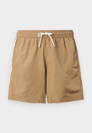 Homme musclé debout pieds nus portant un short beige avec cordon, regardant vers le bas et sur sa gauche sur un fond blanc uni.