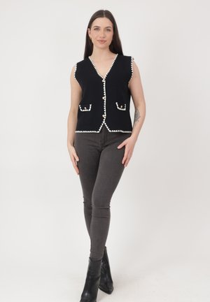 Kvinde står iført sort ærmeløs knapvest med hvid kant og lommer, mørkegrå skinny jeans og sorte ankelstøvler.