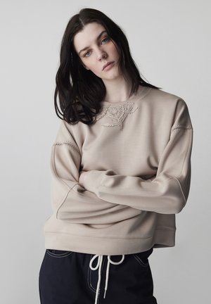 Vrouw met lang donker haar draagt een beige sweatshirt met bloemrijke borduursels en zwarte broek met witte trekkoord, staand tegen een lichte achtergrond.