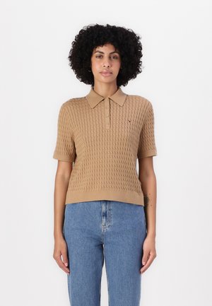 Beige gebreide polo met korte mouwen, voorzien van een gestructureerd patroon, knoopsluiting en een klein logo op de borst. Gecombineerd met blauwe jeans.