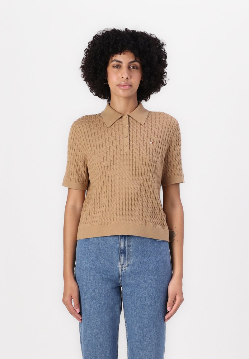 Polo shirt a maglia beige con maniche corte, caratterizzato da un motivo testurizzato, zip e un piccolo logo sul petto. Abbinato a jeans blu.