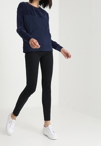 Person som bär en marinblå tröja med långa ärmar, svarta skinny jeans och vita sneakers, stående mot en enfärgad vit bakgrund.
