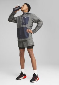 Lätt grå jacka med marinblå detaljer, ficka med dragkedja, matchande grå shorts och svarta sneakers med röda detaljer. Inkluderar vattenflaska.