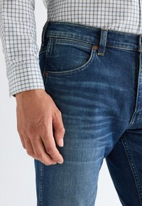 Male wearing blue jeans and a white checkered shirt, with hand resting beside the thigh and visible denim texture.