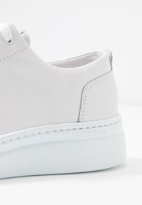 Gros plan de l’arrière et du côté d’une chaussure de sport en cuir blanc avec des coutures blanches et une semelle blanche épaisse sur un fond uni.
