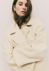 Beige wool-blend coat with a lapel collar and oversized sleeves. The texture appears soft and thick, with a relaxed fit and no visible fastenings.