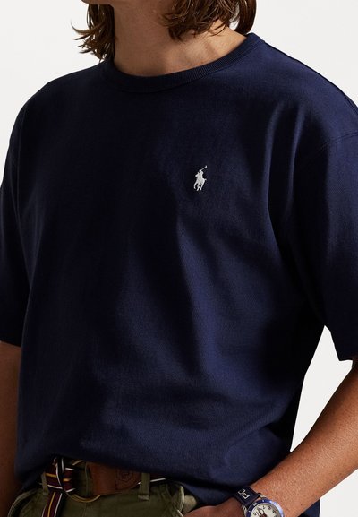 Σκούρο μπλε βαμβακερό μπλουζάκι με κοντά μανίκια και κανονικό λαιμό. Διαθέτει ένα μικρό λευκό κεντημένο λογότυπο στο στήθος. Λείος υφασμάτινος ιστός.