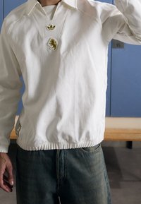 Bílý svetr s výstřihem do V, zdobený zeleným logem a hravým vyšitým motivem zvířete. Elastický pas a manžety.