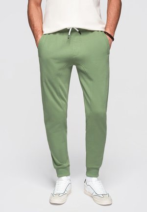 Homme portant un pantalon de jogging vert clair avec des poignets élastiques, un cordon blanc, des baskets blanches et un t-shirt blanc, mains dans les poches.