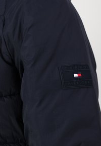 Tamsiai mėlynas pūkinis švarkas su siuvinėtu paviršiumi. Yra stačiakampis paženklinimas su "TOMMY HILFIGER" ir spalvotu logotipu raudonoje, baltos ir mėlynos spalvose.