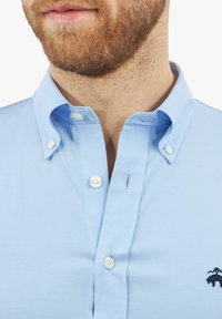 Jasnoniebieska koszula z guzikami o miękkiej fakturze, z białymi guzikami i małym ciemnym logo na klatce piersiowej. Klasyczny krój kołnierza.