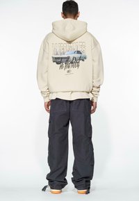 Beige Kapuzenpullover mit Rückendruck eines klassischen Autos, kombiniert mit dunklen Cargo-Hosen mit mehreren Taschen und elastischen Bündchen, getragen mit orange-soled Schuhen.
